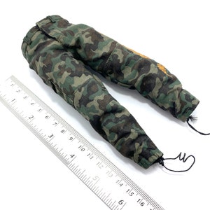 May include: Camouflage trousers with a drawstring at the bottom of each leg. The trousers are green, brown, and black with a small orange patch on the right leg.