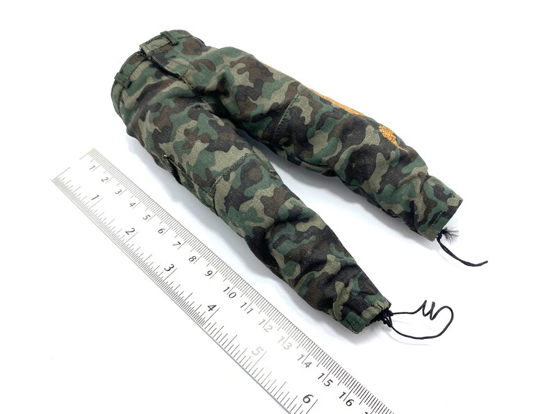 May include: Camouflage trousers with a drawstring at the bottom of each leg. The trousers are green, brown, and black with a small orange patch on the right leg.