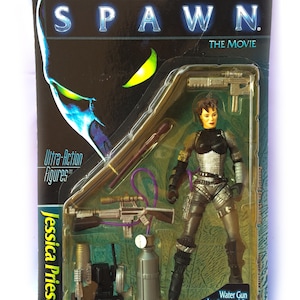 May include: A plastic package containing an action figure of Jessica Priest from the Spawn movie. The figure is wearing a black and silver outfit and holding a gun. The package also includes a water gun, a backpack missile launcher, and a small tank. The package is labeled "Ultra-Action Figures" and "Jessica Priest".