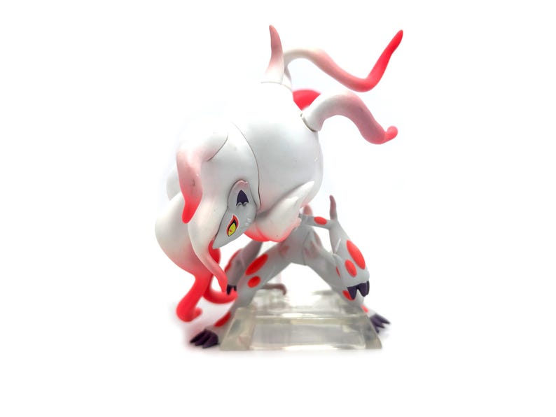Puede incluir: Una figura blanca y gris con detalles en rojo, que representa una criatura en una pose din&aacute;mica. La figura tiene un dise&ntilde;o estilizado con manchas rojas y cuernos curvos. Sobre un fondo blanco.
