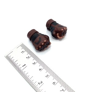 Op de afbeelding: Twee bruine en zwarte leren handschoenen met vingergaten. De handschoenen zijn ongeveer 7,5 cm lang en liggen op een wit oppervlak.