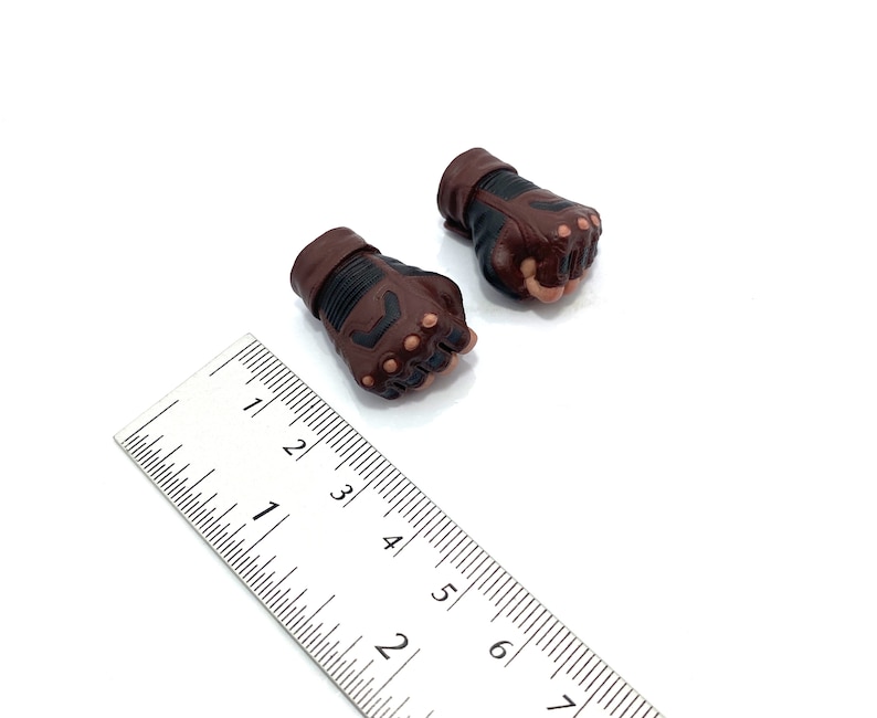 Op de afbeelding: Twee bruine en zwarte leren handschoenen met vingergaten. De handschoenen zijn ongeveer 7,5 cm lang en liggen op een wit oppervlak.