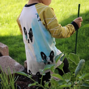 Puede incluir: Una camisa blanca de manga larga con un dise&ntilde;o de acuarela azul y amarillo. La camisa presenta una silueta de perro en blanco y negro con una estrella azul en la espalda.