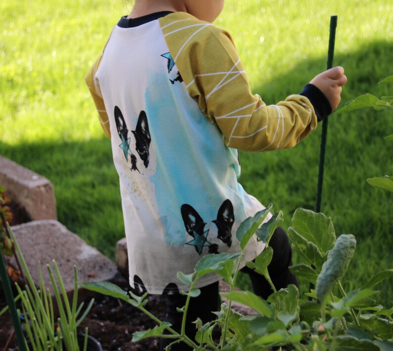 Puede incluir: Una camisa blanca de manga larga con un dise&ntilde;o de acuarela azul y amarillo. La camisa presenta una silueta de perro en blanco y negro con una estrella azul en la espalda.