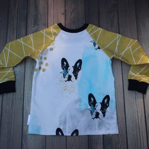 Puede incluir: Una camisa blanca de manga larga con mangas ragl&aacute;n amarillas y negras. La camisa presenta un dise&ntilde;o de acuarela con dos ilustraciones de Bulldog Franc&eacute;s en blanco y negro, cada una con una estrella dorada en el ojo.