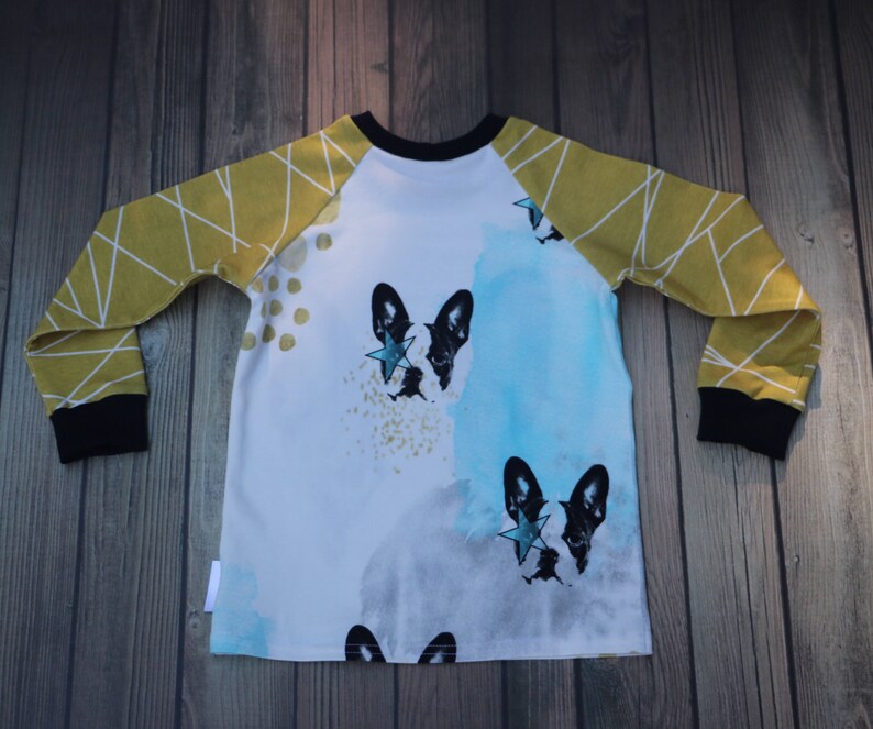 Puede incluir: Una camisa blanca de manga larga con mangas ragl&aacute;n amarillas y negras. La camisa presenta un dise&ntilde;o de acuarela con dos ilustraciones de Bulldog Franc&eacute;s en blanco y negro, cada una con una estrella dorada en el ojo.