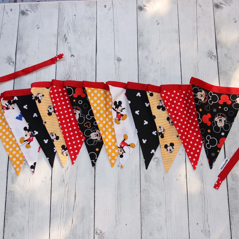 May include: A Mickey Mouse themed party banner with 10 red, yellow, and white pennants featuring Mickey Mouse in different poses. The pennants are strung together with red ribbon.
