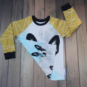 Puede incluir: Camiseta blanca de manga larga con un gr&aacute;fico azul y blanco de la cara de un perro con una estrella. Las mangas son amarillas con patrones geom&eacute;tricos blancos.