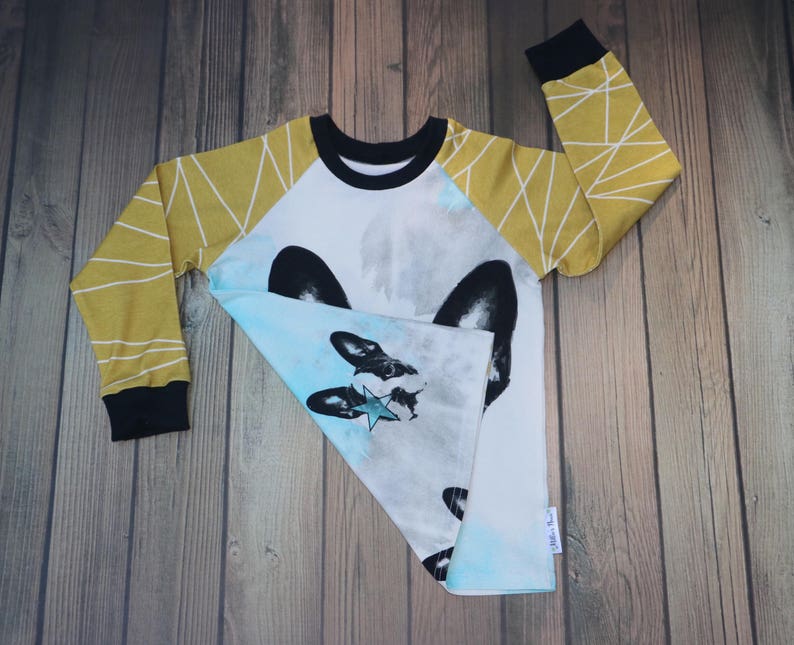 Puede incluir: Camiseta blanca de manga larga con un gr&aacute;fico azul y blanco de la cara de un perro con una estrella. Las mangas son amarillas con patrones geom&eacute;tricos blancos.