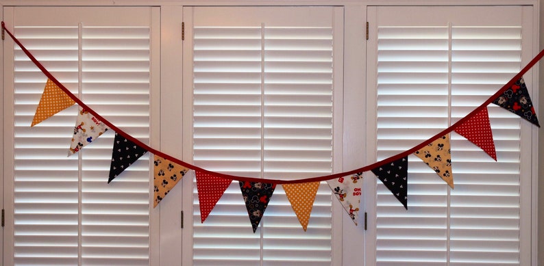 May include: A red, yellow, and black fabric banner with a Mickey Mouse theme. The banner is made of triangular flags with different patterns, including polka dots, stripes, and Mickey Mouse characters.
