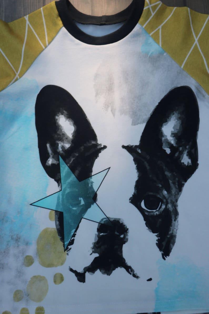 Puede incluir: Una camiseta blanca con un dise&ntilde;o de acuarela de un bulldog franc&eacute;s con una estrella azul sobre el ojo. El fondo es un dise&ntilde;o de acuarela azul claro y amarillo.
