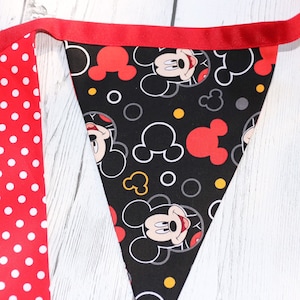 May include: A black and red fabric banner with a Mickey Mouse pattern. The banner is made of a soft, cotton fabric and features a repeating pattern of Mickey Mouse's head, ears, and face. The banner is trimmed with a red ribbon.