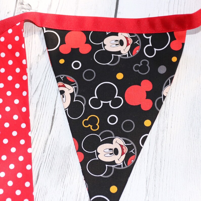 May include: A black and red fabric banner with a Mickey Mouse pattern. The banner is made of a soft, cotton fabric and features a repeating pattern of Mickey Mouse's head, ears, and face. The banner is trimmed with a red ribbon.