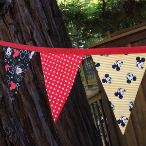 May include: A red, white, and yellow bunting with a Mickey Mouse theme. The bunting is made of fabric and has a red trim. The bunting is hanging from a tree branch.
