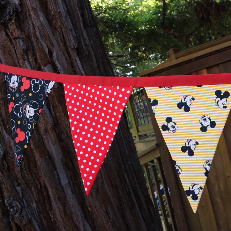 May include: A red, white, and yellow bunting with a Mickey Mouse theme. The bunting is made of fabric and has a red trim. The bunting is hanging from a tree branch.