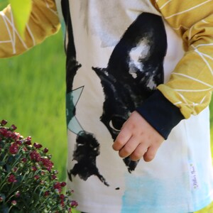 Puede incluir: Una camisa blanca de manga larga con un estampado gr&aacute;fico en blanco y negro de la cara de un perro. La camisa tiene mangas a rayas amarillas y blancas con un pu&ntilde;o negro. La camisa tiene una peque&ntilde;a etiqueta blanca con el texto "Little's Place" en la esquina inferior derecha.
