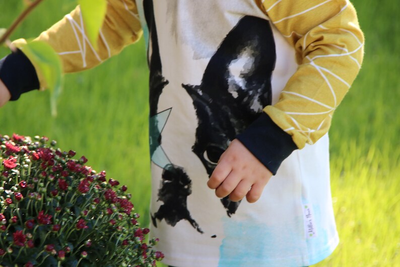 Puede incluir: Una camisa blanca de manga larga con un estampado gr&aacute;fico en blanco y negro de la cara de un perro. La camisa tiene mangas a rayas amarillas y blancas con un pu&ntilde;o negro. La camisa tiene una peque&ntilde;a etiqueta blanca con el texto "Little's Place" en la esquina inferior derecha.