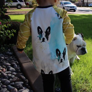 Puede incluir: Un ni&ntilde;o peque&ntilde;o con una camisa blanca de manga larga con un dise&ntilde;o de acuarela azul y amarillo que presenta un bulldog franc&eacute;s blanco y negro con una estrella azul en la cabeza. La camisa tiene rayas amarillas en las mangas. El ni&ntilde;o est&aacute; de pie junto a un bulldog blanco.