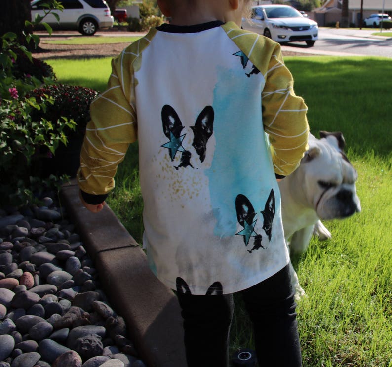 Puede incluir: Un ni&ntilde;o peque&ntilde;o con una camisa blanca de manga larga con un dise&ntilde;o de acuarela azul y amarillo que presenta un bulldog franc&eacute;s blanco y negro con una estrella azul en la cabeza. La camisa tiene rayas amarillas en las mangas. El ni&ntilde;o est&aacute; de pie junto a un bulldog blanco.