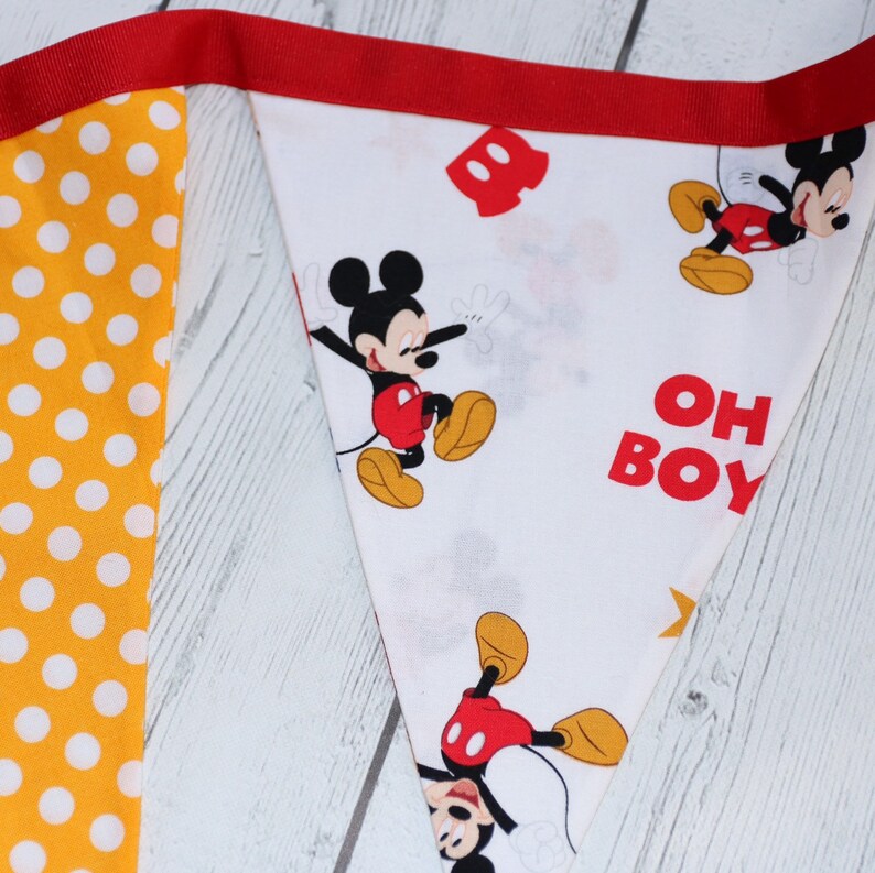 May include: A white fabric banner with a red border and a Mickey Mouse theme. The banner features a repeating pattern of Mickey Mouse in various poses, along with the text "OH BOY". The banner is decorated with yellow polka dots.