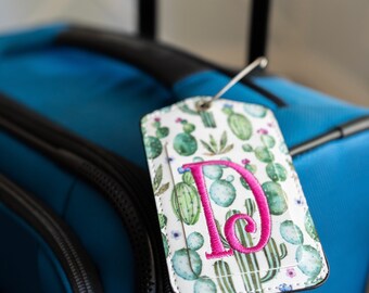 以下が含まれることがあります： 緑色のサボテン柄とピンク色の刺繍で「D」の文字が施された白い荷物タグです。