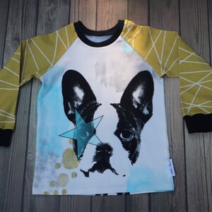 Puede incluir: Una camisa blanca de manga larga con un patr&oacute;n geom&eacute;trico amarillo mostaza y blanco en las mangas. La camisa presenta una ilustraci&oacute;n en blanco y negro de un Bulldog Franc&eacute;s con una estrella azul sobre un ojo.