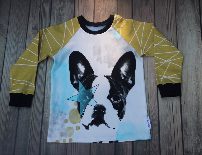 Puede incluir: Una camisa blanca de manga larga con un patr&oacute;n geom&eacute;trico amarillo mostaza y blanco en las mangas. La camisa presenta una ilustraci&oacute;n en blanco y negro de un Bulldog Franc&eacute;s con una estrella azul sobre un ojo.