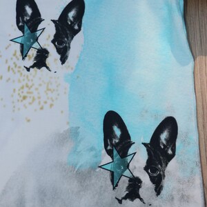 Puede incluir: Una camiseta blanca con un fondo de acuarela azul y gris. Dos caras de bulldog franc&eacute;s en blanco y negro est&aacute;n impresas en la camiseta, cada una con una estrella azul sobre un ojo.