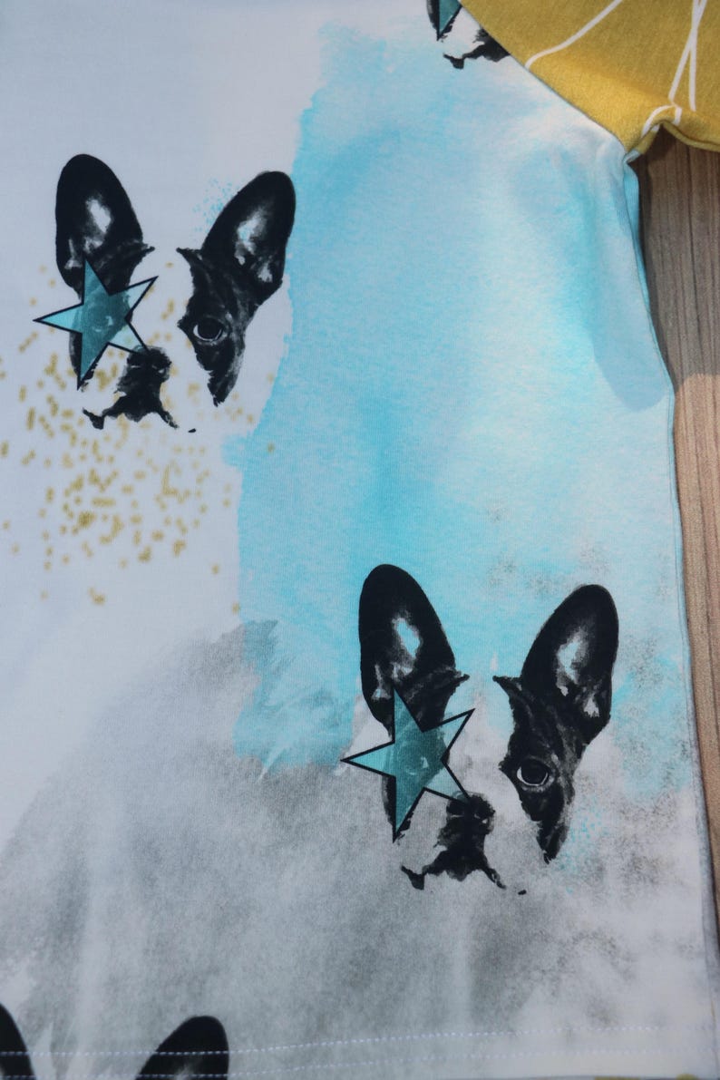 Puede incluir: Una camiseta blanca con un fondo de acuarela azul y gris. Dos caras de bulldog franc&eacute;s en blanco y negro est&aacute;n impresas en la camiseta, cada una con una estrella azul sobre un ojo.
