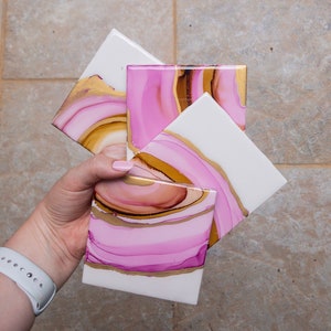 Può includere: Set di quattro sottobicchieri quadrati con disegni astratti a spirale rosa, oro e marrone. I sottobicchieri sono in ceramica e hanno una finitura lucida.