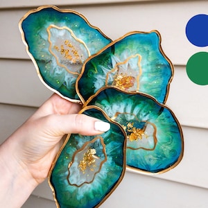 Peut inclure: Quatre sous-verres en agate bleu-vert avec des accents en feuille d'or et un bord doré. Les sous-verres sont empilés et tenus par une main.