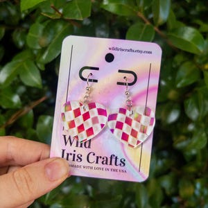 May include: A pair of heart-shaped earrings with a checkered pattern in red and white. The earrings are made of clear resin and have silver hooks. The earrings are displayed on a card with the text "Wild Iris Crafts" and "Made with love in the USA".