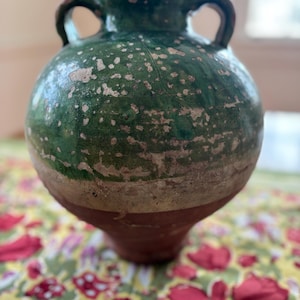 May include: A green and brown ceramic vase with two handles. The vase has a rounded body and a flared base. The green glaze is speckled with white, and the vase has a rustic, aged appearance. The vase is sitting on a floral patterned surface.