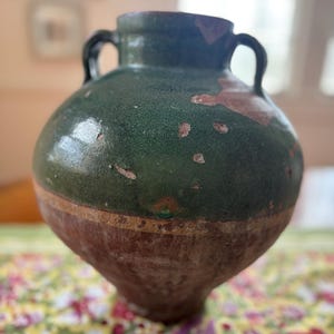May include: A large, antique, two-handled ceramic jar with a rounded body. The upper portion is glazed in a dark green, while the lower half is a reddish-brown. The jar shows signs of age with chipped areas, adding to its vintage appeal.