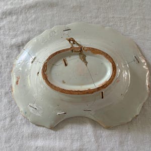 Puede incluir: Un plato de cerámica blanco con un borde festoneado y una grieta que atraviesa el centro. El plato tiene un borde marrón y un agujero en el centro para colgar.