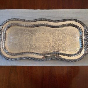May include: A rectangular silver-toned serving tray featuring ornate, embossed floral and scrollwork patterns across the center surface. The tray has a scalloped, raised border with a rope-like edge and two decorative handles with intricate detailing.