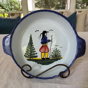 Può includere: Un piatto per torte in ceramica blu e bianca con un bordo blu e due manici. Il piatto presenta un disegno dipinto a mano di una persona con una giacca blu e una gonna rossa in piedi in un bosco.