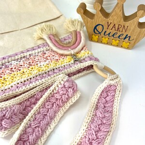 May include: A crocheted bag with a pink and white braided strap, a rainbow-coloured yarn tassel, and a wooden sign that says "Yarn Queen".