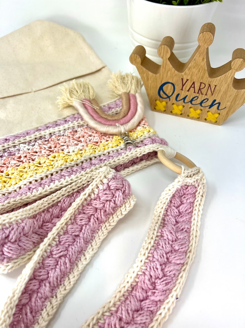 May include: A crocheted bag with a pink and white braided strap, a rainbow-coloured yarn tassel, and a wooden sign that says "Yarn Queen".