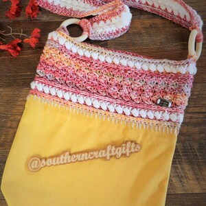 May include: A yellow crocheted tote bag with a pink, white and orange crocheted strap. The bag has a wooden ring on the strap and a wooden tag that says "@southerncraftgifts".