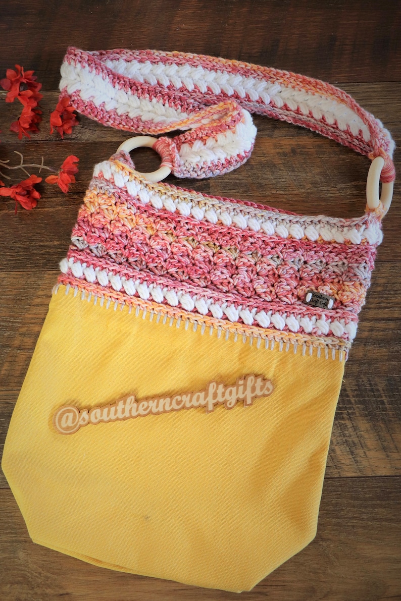 May include: A yellow crocheted tote bag with a pink, white and orange crocheted strap. The bag has a wooden ring on the strap and a wooden tag that says "@southerncraftgifts".
