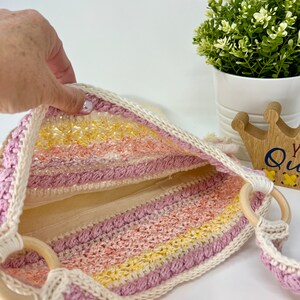 May include: A pink, yellow and white striped crochet tote bag with wooden handles. The bag is lined with a white fabric.