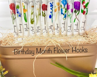 May include: Twelve white ceramic hooks with different pressed flowers and the month of the year written on each hook. The hooks are displayed in a brown metal container with the text "Birthday Month Flower Hooks" and "@HookedbyHoltby" on the container.