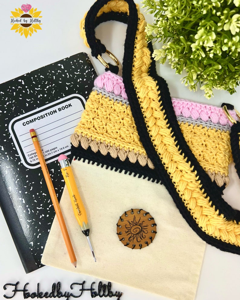 May include: A yellow, black, pink and grey crocheted pencil bag with a black strap. The bag is sitting on a white surface with a composition book, a pencil and a crochet hook. The bag has a wooden sun charm attached to it.