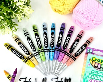 Puede incluir: Un juego de ocho agujas de crochet, cada una con forma de crayón y etiquetada con una letra y un tamaño en milímetros. Las agujas están dispuestas en semicírculo, con la aguja más pequeña a la izquierda y la más grande a la derecha. Las agujas están rodeadas de hilo y crayones de colores.