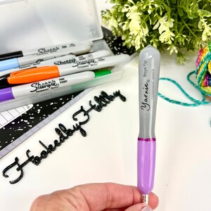 Pu&ograve; includere: Un pennarello permanente Yarnie Boye Tip di colore viola e grigio con una punta argentata. Il pennarello &egrave; tenuto in una mano su uno sfondo bianco. Il testo "Yarnie Boye Tip" e "Permanent Marker" &egrave; stampato sul pennarello.