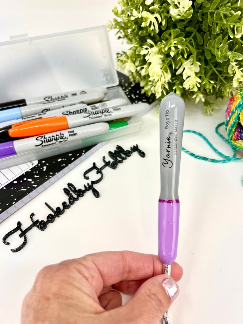 Pu&ograve; includere: Un pennarello permanente Yarnie Boye Tip di colore viola e grigio con una punta argentata. Il pennarello &egrave; tenuto in una mano su uno sfondo bianco. Il testo "Yarnie Boye Tip" e "Permanent Marker" &egrave; stampato sul pennarello.
