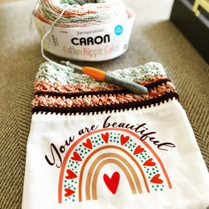 May include: A white crocheted bag with a brown crocheted border. The bag features a rainbow design with red, orange, yellow, green, and blue stripes. The rainbow has a red heart in the centre and is surrounded by green and red hearts. The bag has the text "You are beautiful" printed on it.