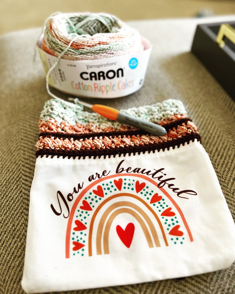 May include: A white crocheted bag with a brown crocheted border. The bag features a rainbow design with red, orange, yellow, green, and blue stripes. The rainbow has a red heart in the centre and is surrounded by green and red hearts. The bag has the text "You are beautiful" printed on it.