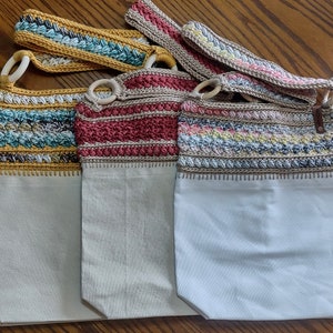 May include: Three crocheted tote bags with different coloured patterns. The bags are made with a white canvas base and have a crocheted strap.